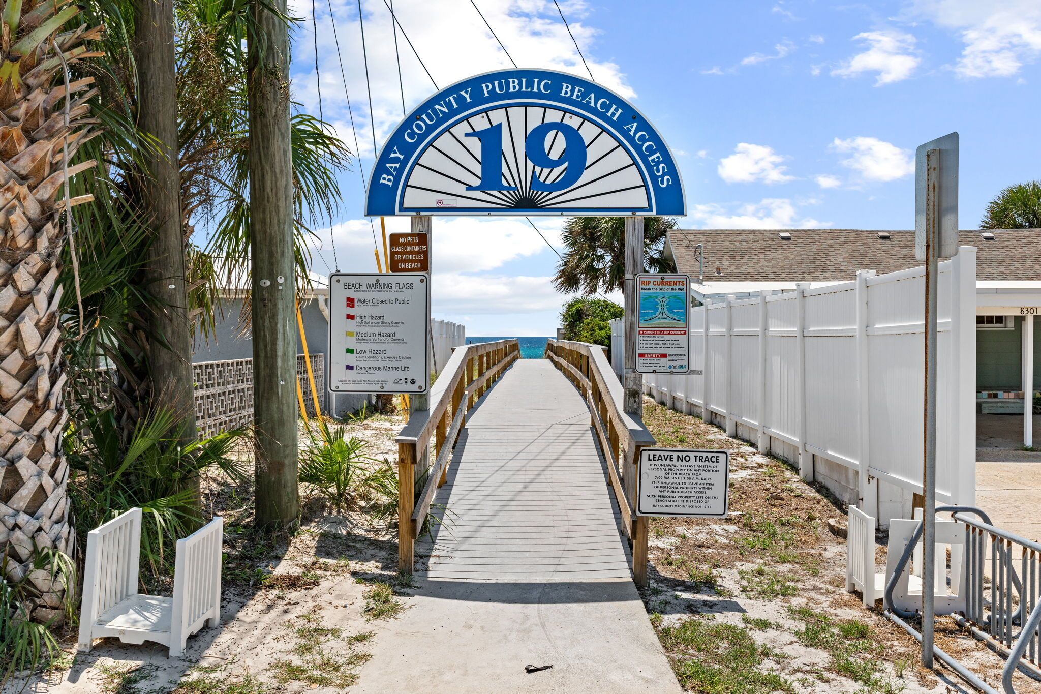 A beach entrance with a sign indicating Bay County Public Beach Access, a warning flag sign, and a 'Leave No Trace' sign.