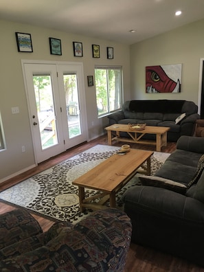 A living room with a sofa, a coffee table, and a patterned rug. There are framed pictures on the wall and a window with a view of trees. - White River Riverside Trout Getaway, 3BR, Sleeps 8, Riverside Pavillion (Norfork)