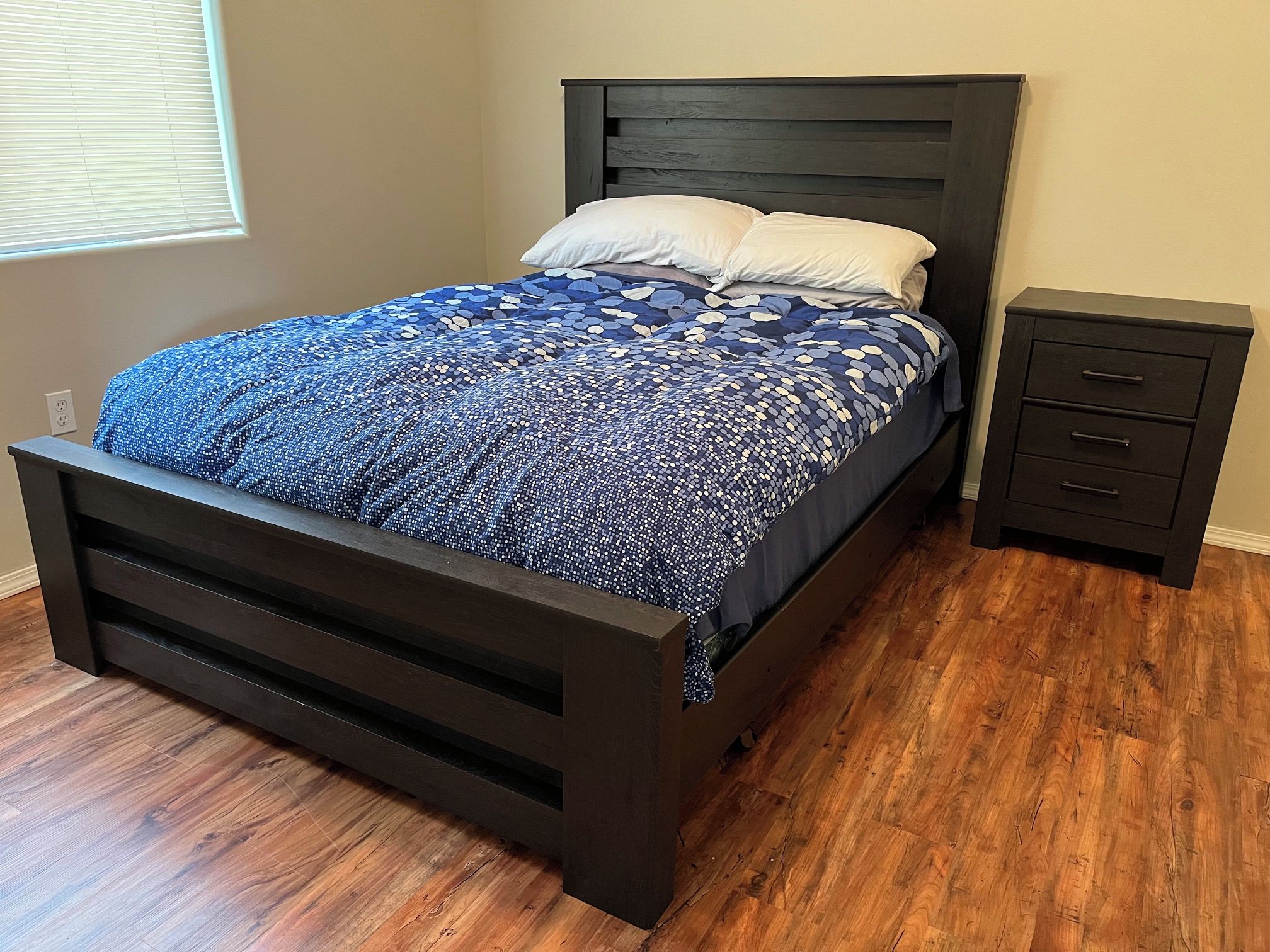 A bed with a dark headboard and a patterned bedspread, a nightstand with drawers, and a window with blinds.