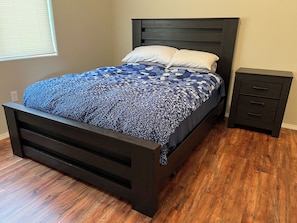 A bed with a dark headboard and a patterned bedspread, a nightstand with drawers, and a window with blinds. - White River Riverside Trout Getaway, 3BR, Sleeps 8, Riverside Pavillion (Norfork)