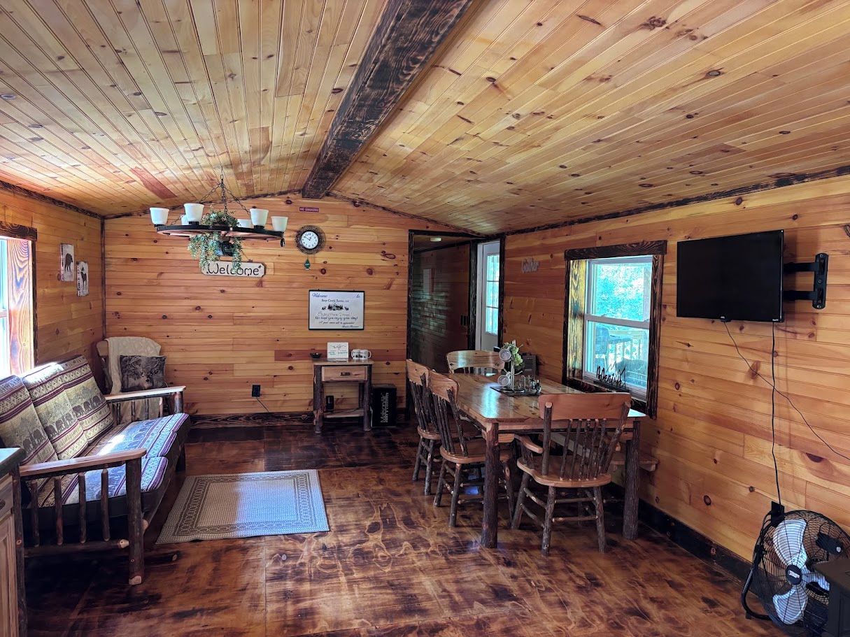 A wooden cabin interior with a sofa, dining table, chairs, a clock, and a television.