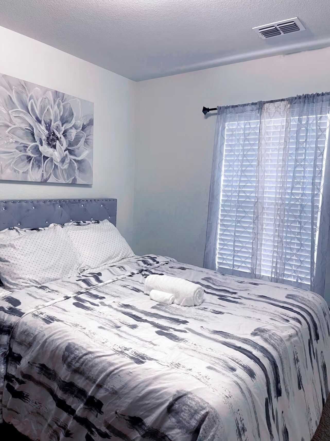 Une chambre à coucher avec un lit, une grande œuvre d’art florale accrochée au mur et une fenêtre avec des stores.