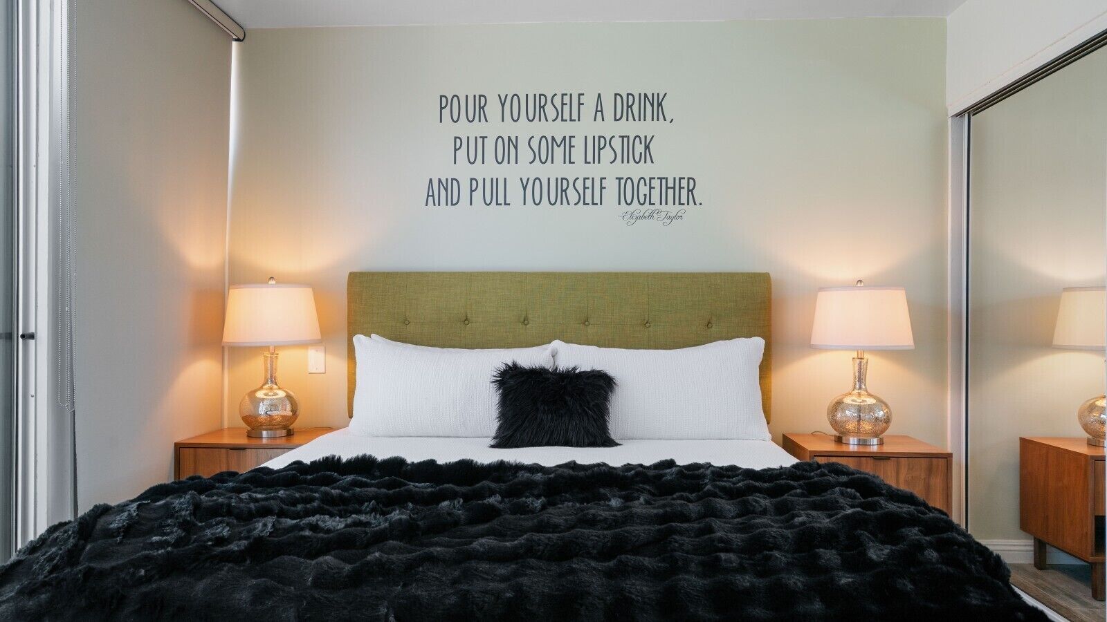 A bedroom with a bed, two bedside lamps, a quote on the wall, and a black furry throw blanket.