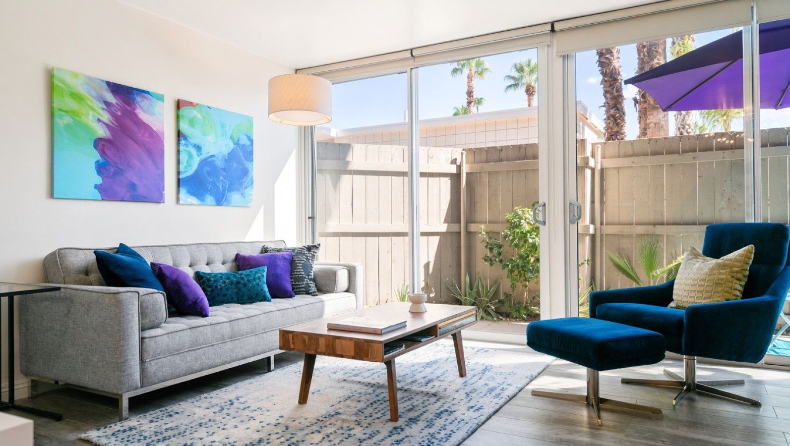 A living room with a grey sofa, blue armchair, and a coffee table. There are abstract paintings on the wall and a view of a wooden fence and palm trees outside.