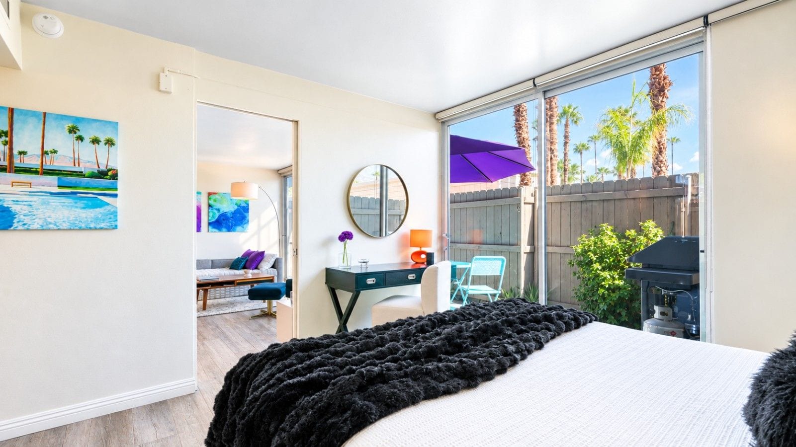 A bedroom with a bed, a desk, a chair, a mirror, and a view of the outside.