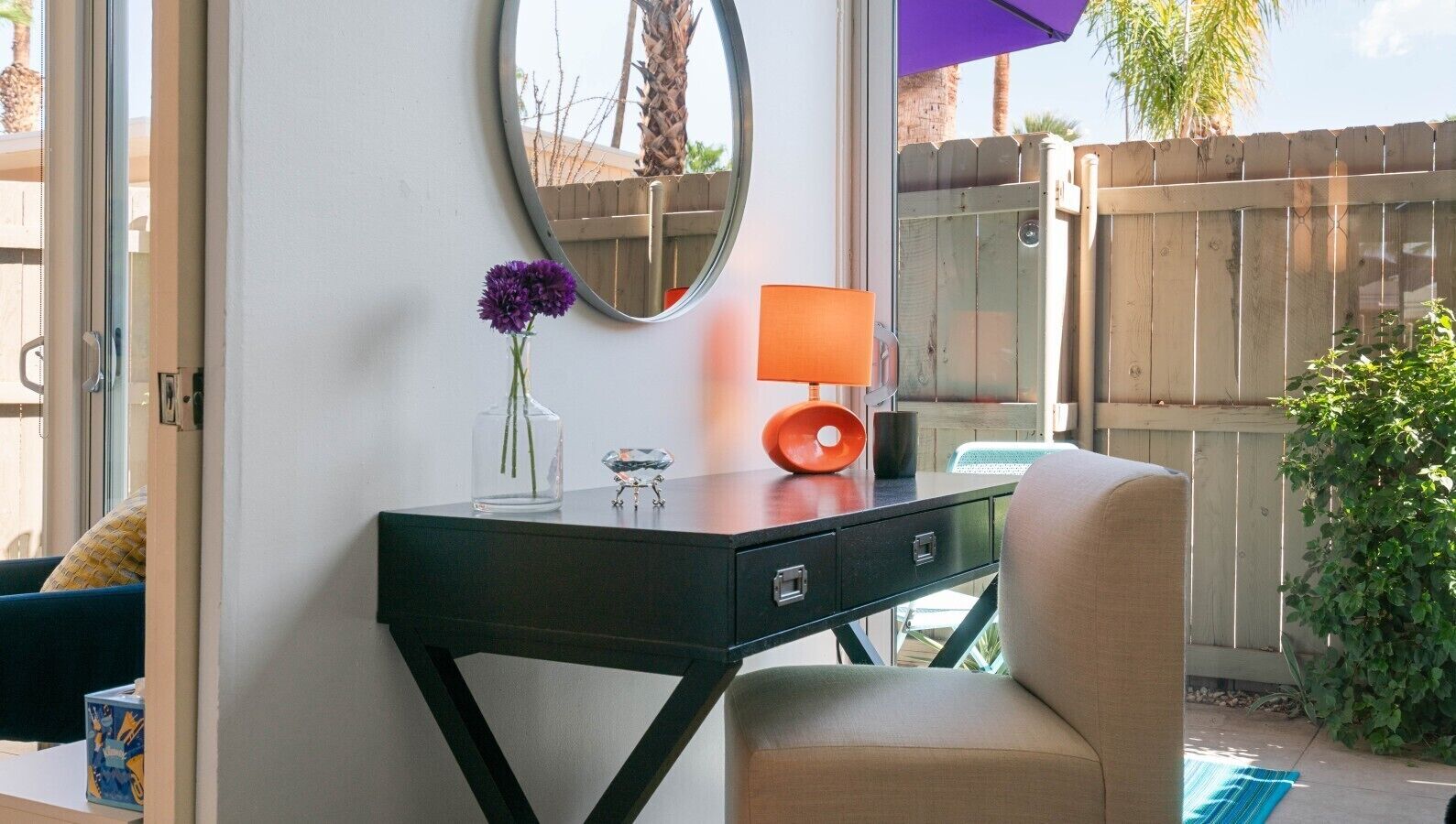Una habitación con escritorio, silla, espejo, un jarrón con flores y una lámpara.