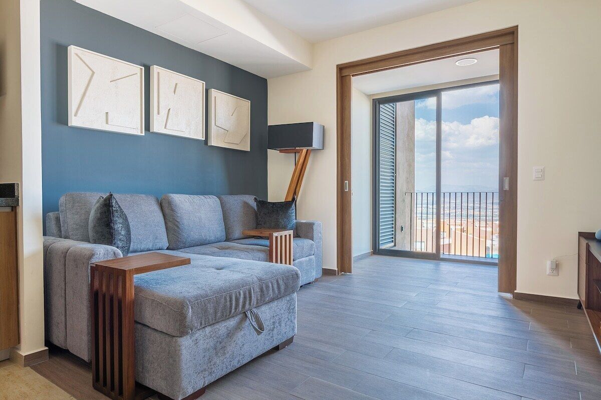 A modern living room with a grey sofa, a wooden side table, and a floor lamp. There is a large window with a view of the outdoors.