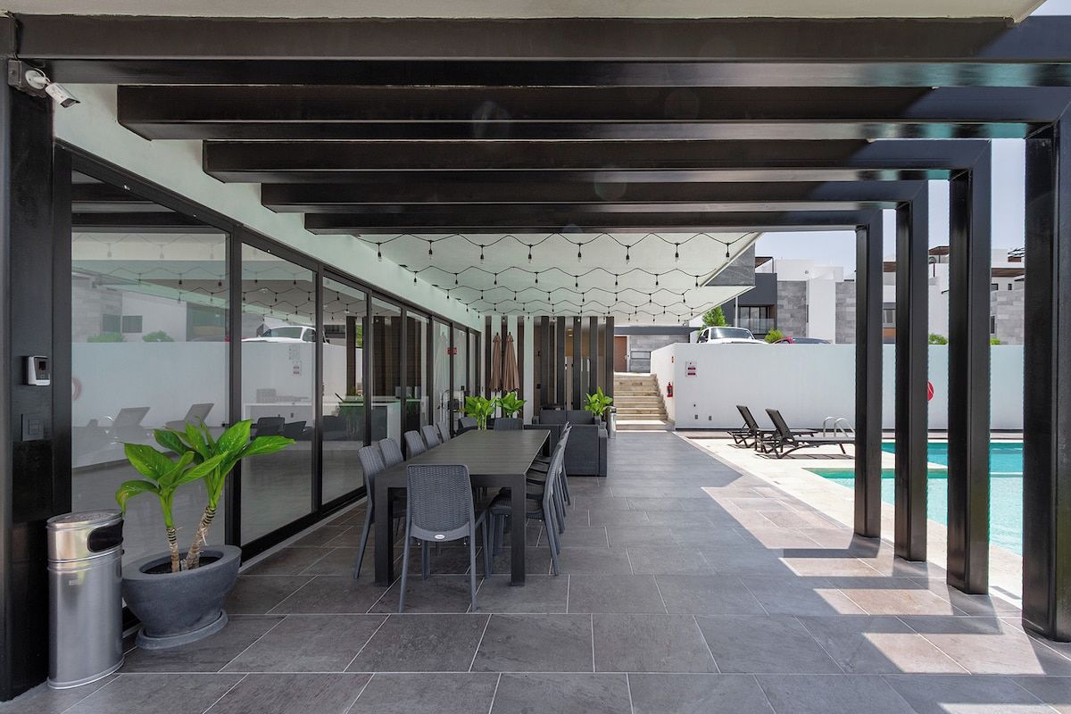 Área externa coberta com mesa, cadeiras, plantas em vasos e piscina ao fundo.