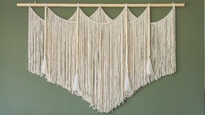 Una stanza con un divano rivestito di grigio, un arazzo in macramè e diversi cuscini decorati.