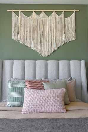 Een kamer met een grijs gestoffeerde bank, een macramé wanddecoratie en verschillende gekleurde kussens.