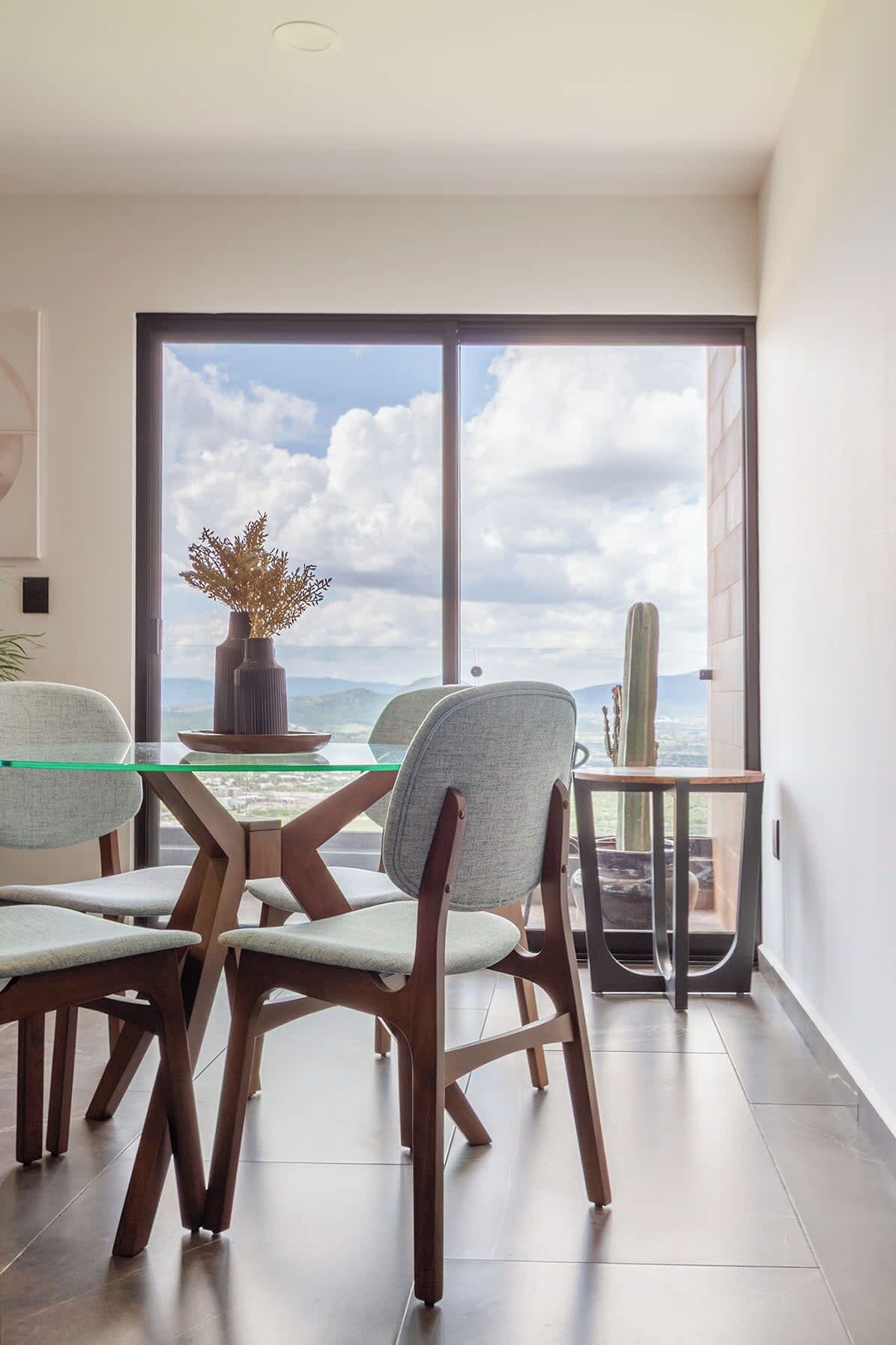A modern dining area with a glass table, wooden chairs, and a view of the sky and mountains.