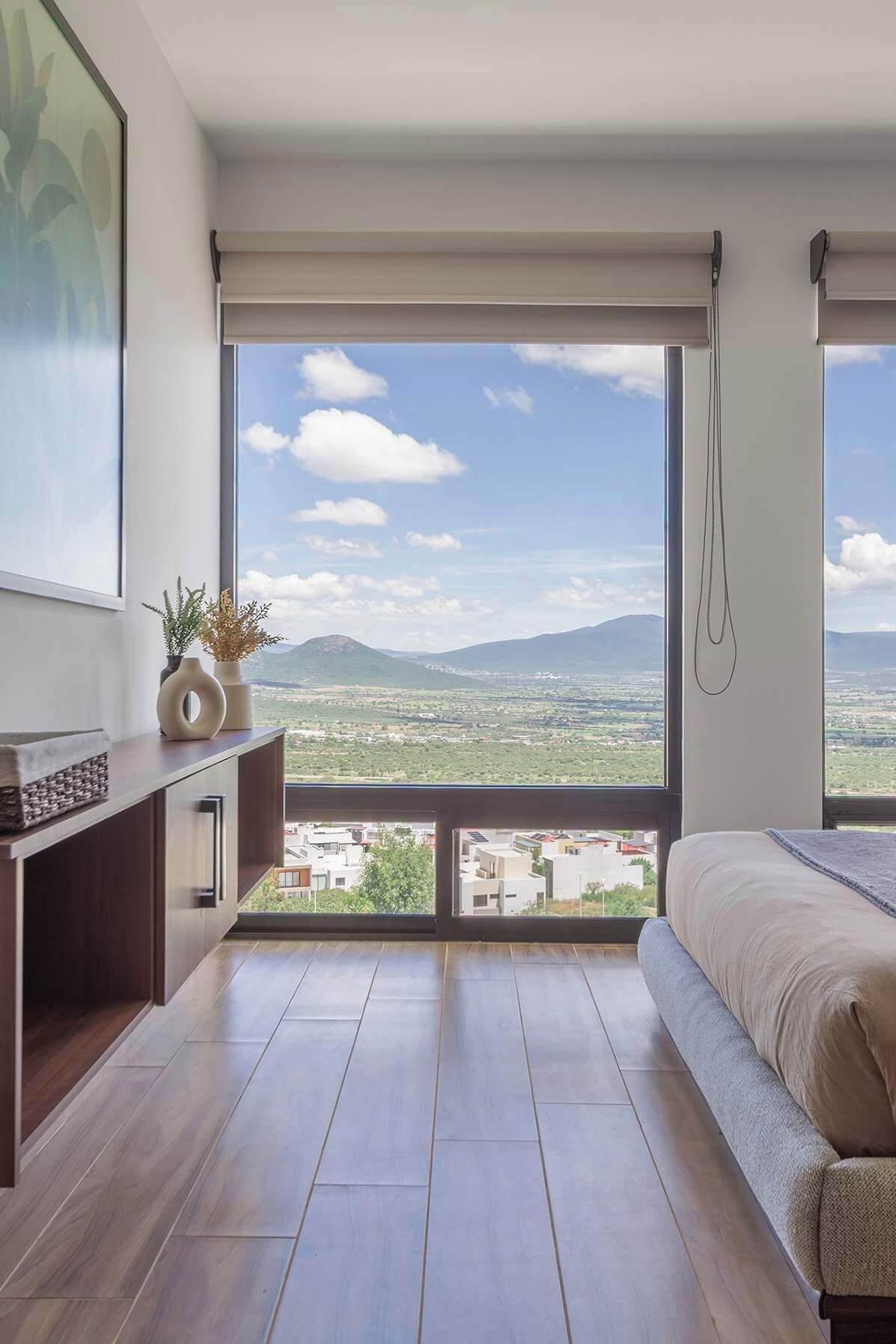Een slaapkamer met een groot raam, een bed, een bureau en uitzicht op een stadsbeeld.