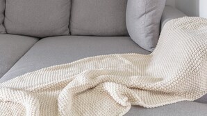 Un divano angolare grigio con una coperta a maglia bianca.