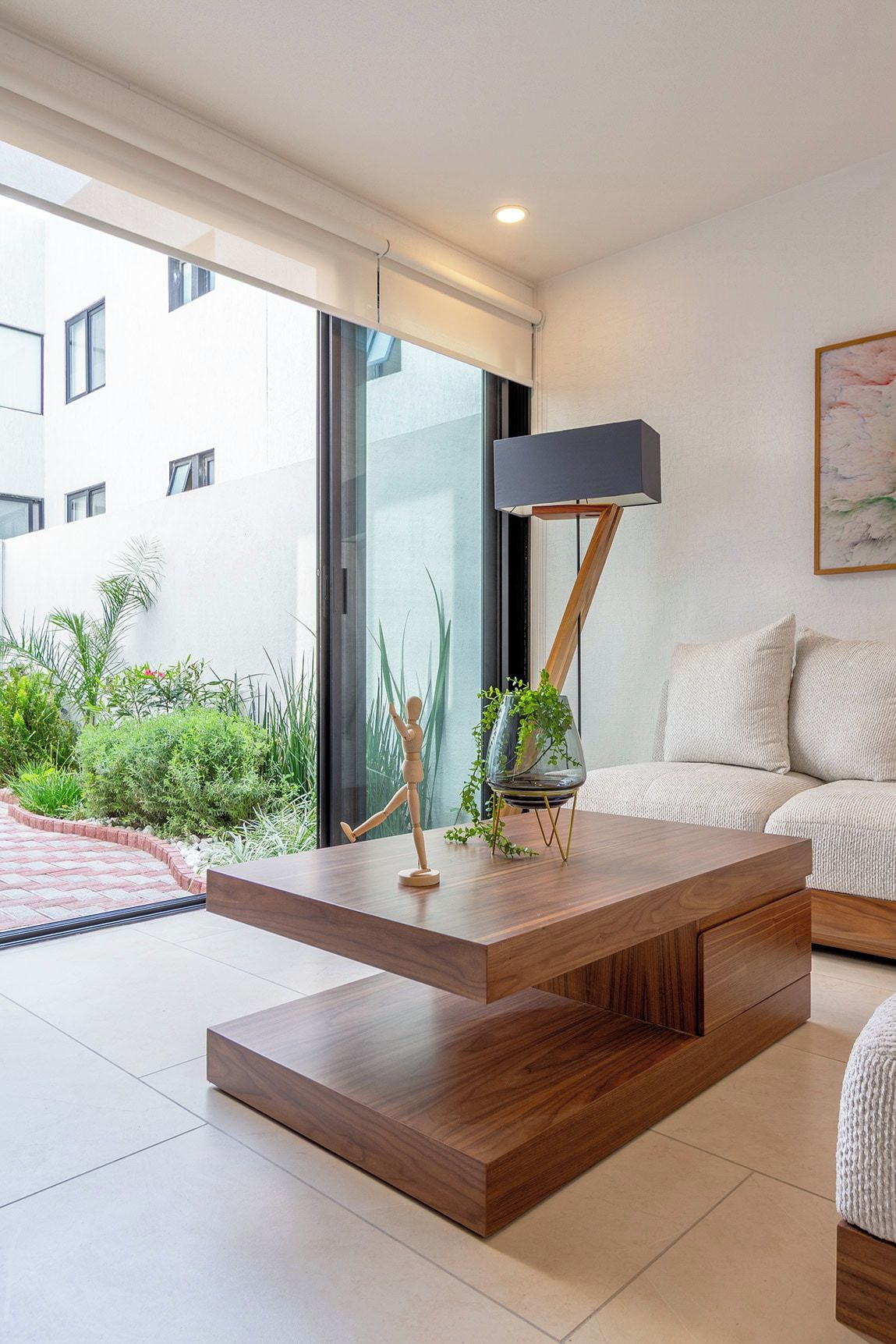 A modern living room with a wooden coffee table, a floor lamp, a sofa, and a view of a building through large windows.