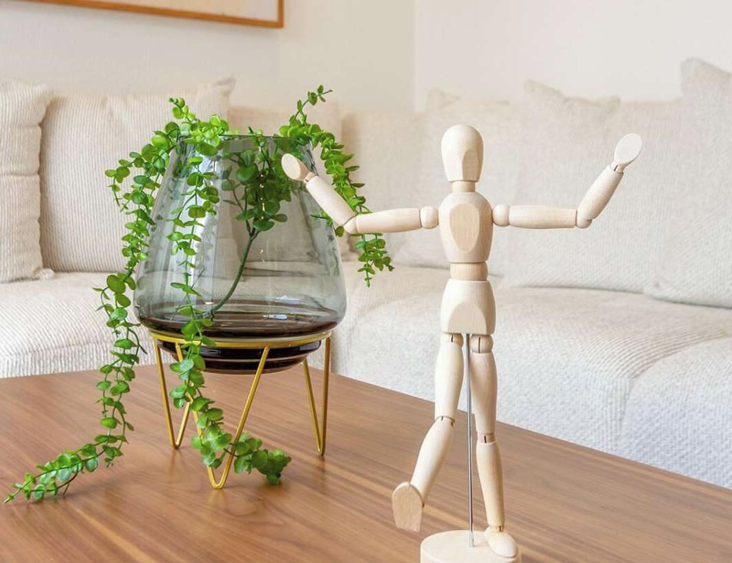 A wooden mannequin on a table with a glass vase containing green plants on a stand.