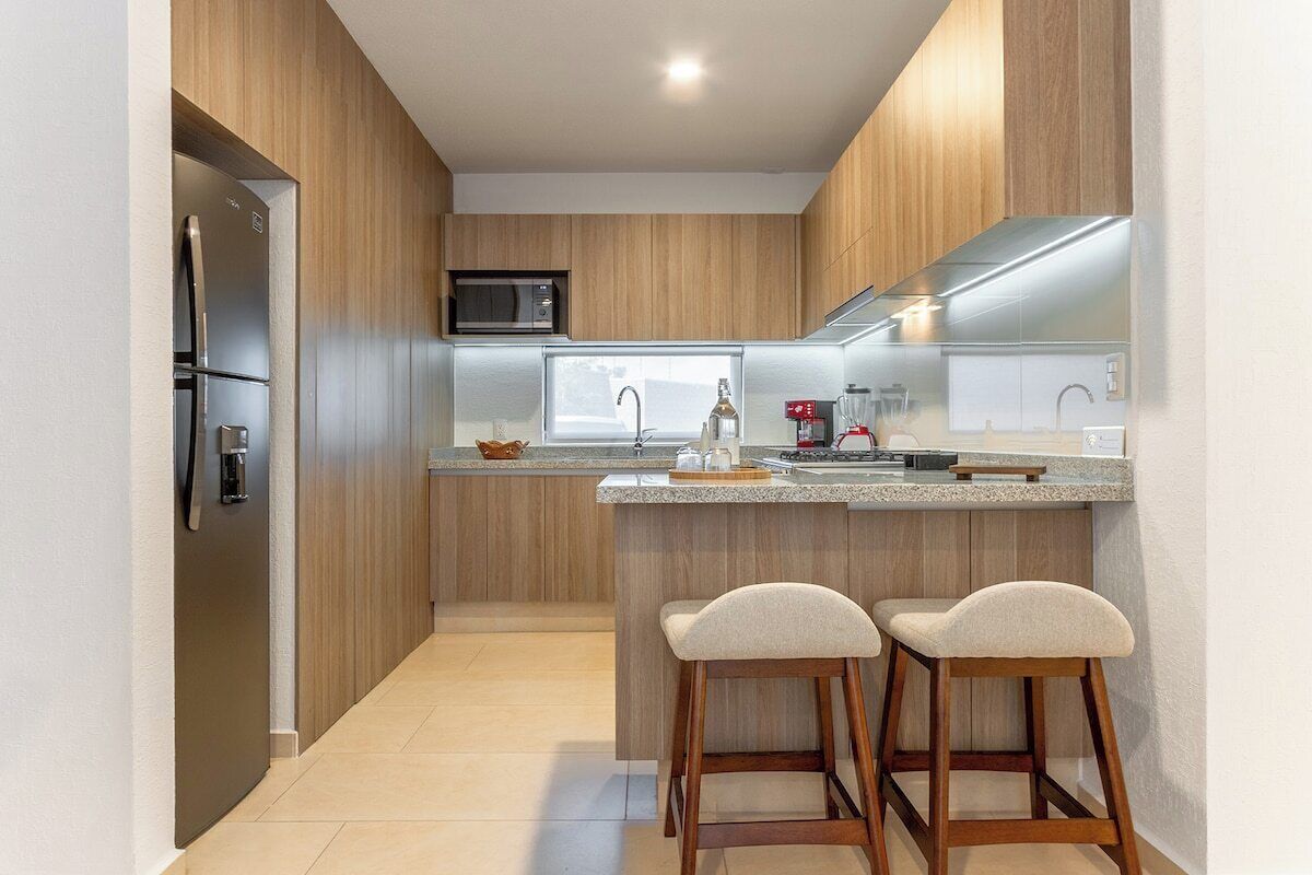 A modern kitchen with wooden cabinets, a built-in microwave, a stainless steel refrigerator, a granite countertop, and two bar stools.