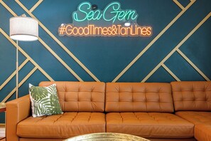 Een kamer met een neonbord waarop staat 'Sea Gem #GoodTimes&TanLines' en een bruine leren bank.