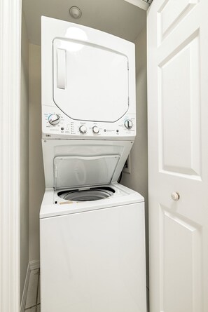 Un appareil compact, blanc, combinant une machine à laver et un sèche-linge à chargement frontal, installé dans un petit espace buanderie, avec une porte sur le côté droit.