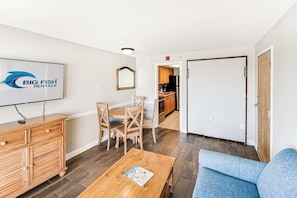 A furnished living room with a wooden coffee table, a blue sofa, a dining area with wooden chairs, a kitchen visible through an open door, and a flat-screen TV mounted on the wall. - Amazing Oceanfront View  Condo with Pool & Hot tub (Myrtle Beach)