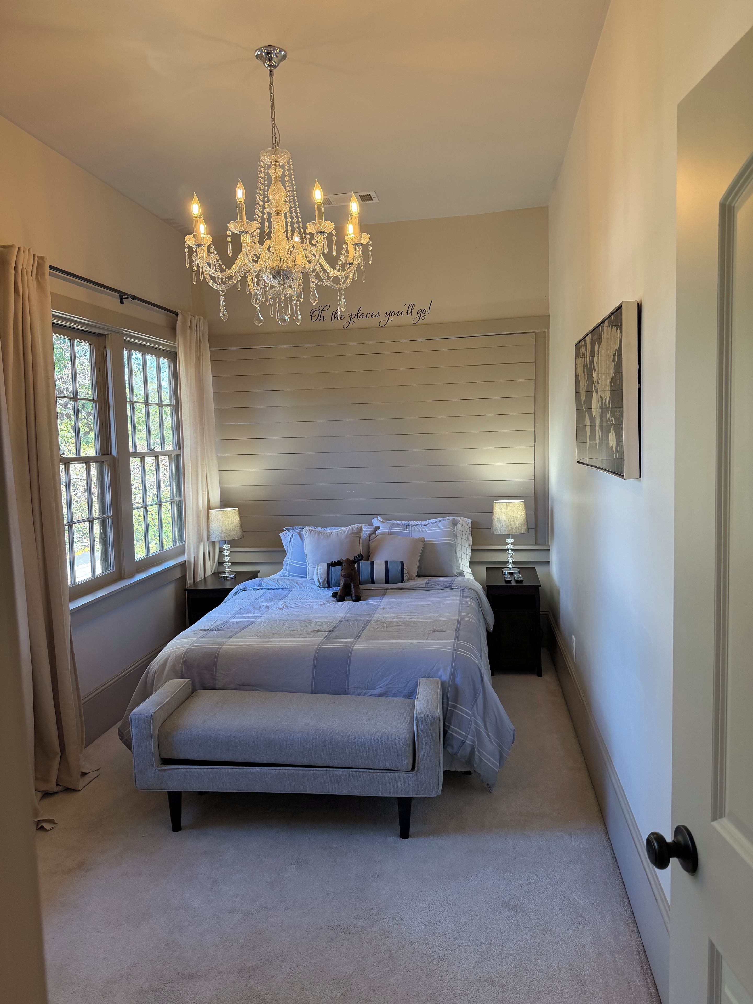 A bedroom with a large bed, a chandelier, a bedside table with a lamp, and a window with curtains.