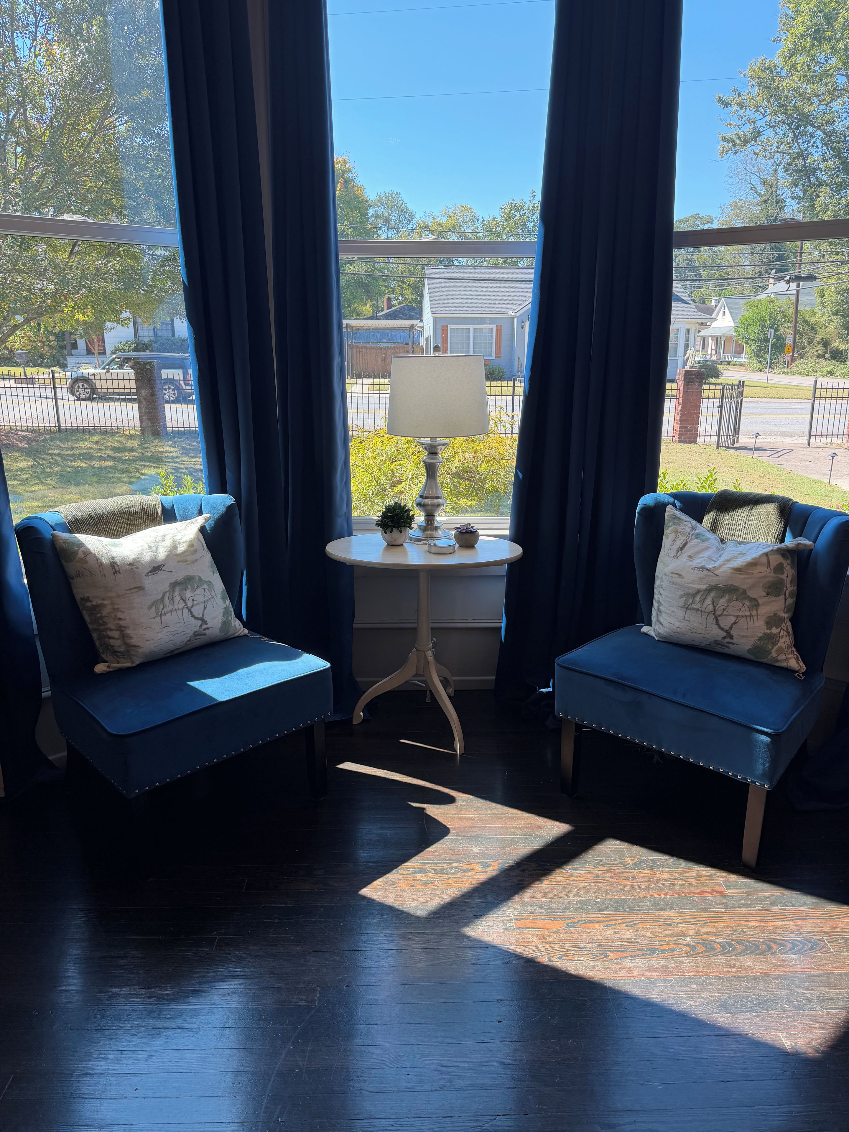 Una habitación con dos sillones azules, una mesita con lámpara, y vistas a una zona residencial a través de amplios ventanales.