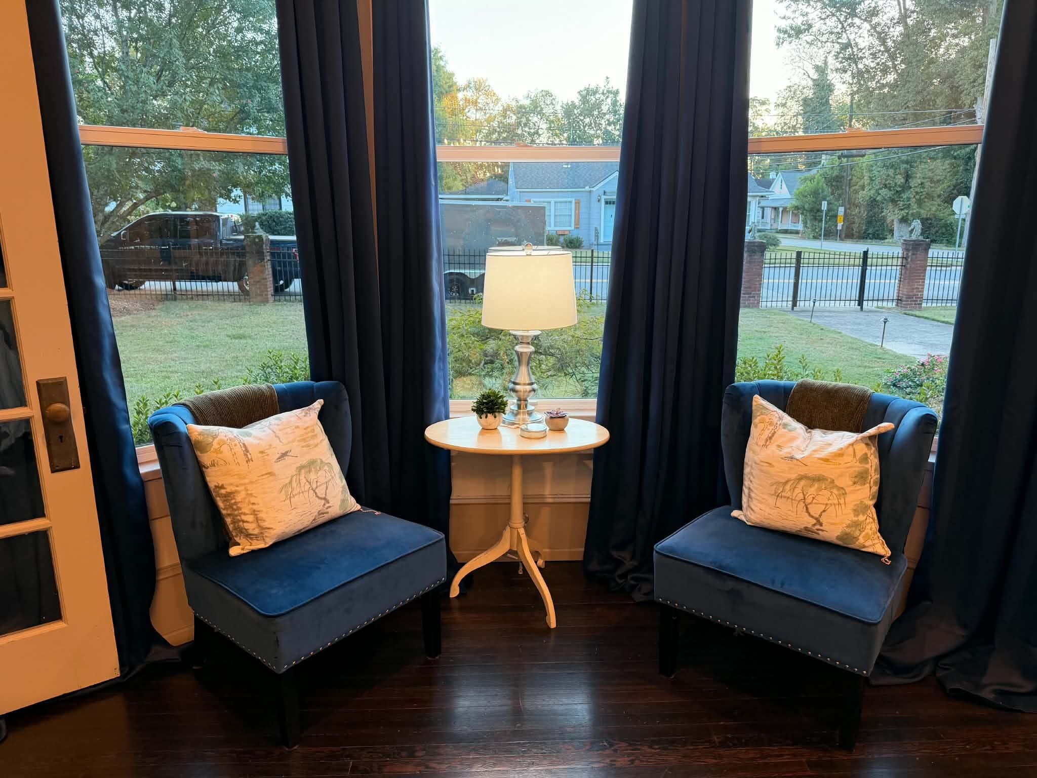 Une pièce avec deux fauteuils bleus, une petite table ronde avec une lampe, et une vue sur un quartier résidentiel grâce à de grandes fenêtres.