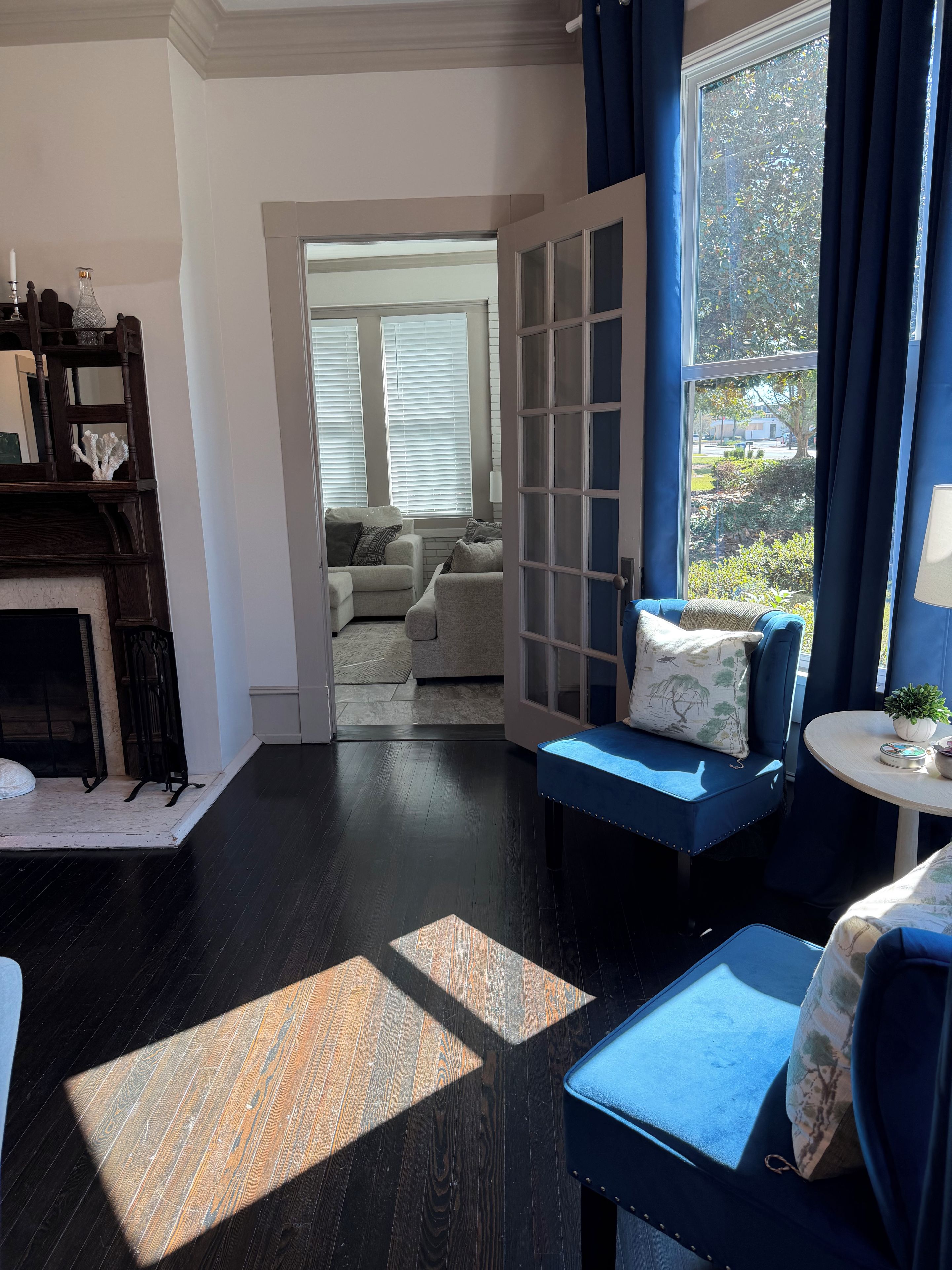 Una habitación con suelo de madera oscura, un sofá azul con cojines, una pequeña mesa redonda con una planta y vistas al exterior a través de una ventana con cortinas.