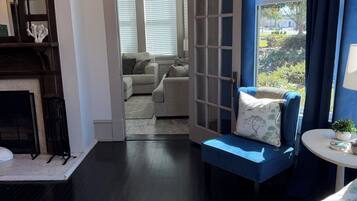 Una habitación con suelo de madera oscura, un sofá azul con cojines, una pequeña mesa redonda con una planta y vistas al exterior a través de una ventana con cortinas.