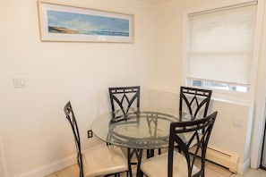 A dining area with a glass table surrounded by four chairs, a framed picture on the wall, and a window with blinds. - Newly Renovated Spacious Stay Near Casino, Boston, Cambridge, & Somerville (Everett)