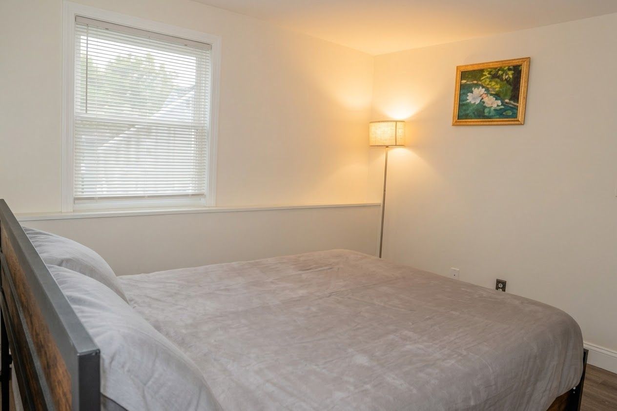 Ein Schlafzimmer mit einem Bett, einem Fenster mit Jalousien, einer Wandlampe und einem gerahmten Bild.