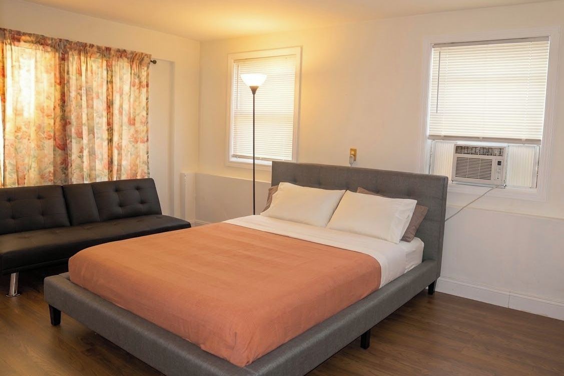 A bedroom with a bed, a sofa, a standing lamp, and an air conditioning unit.