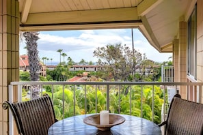 A balcony with a table and chairs, overlooking a lush garden and buildings. - Haleakala Shores | 4BR Total | Sleeps 12 (Kihei)
