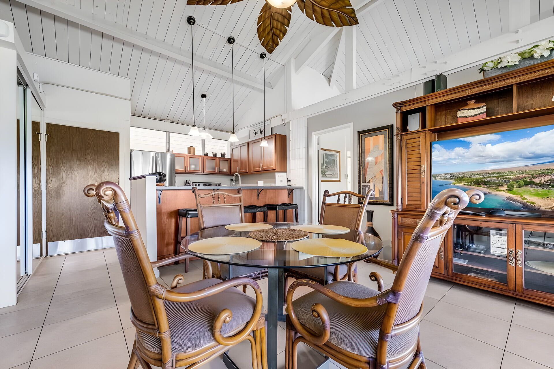 A dining area with a round glass table and four chairs, a kitchen with wooden cabinets, and a large TV displaying a scenic view.