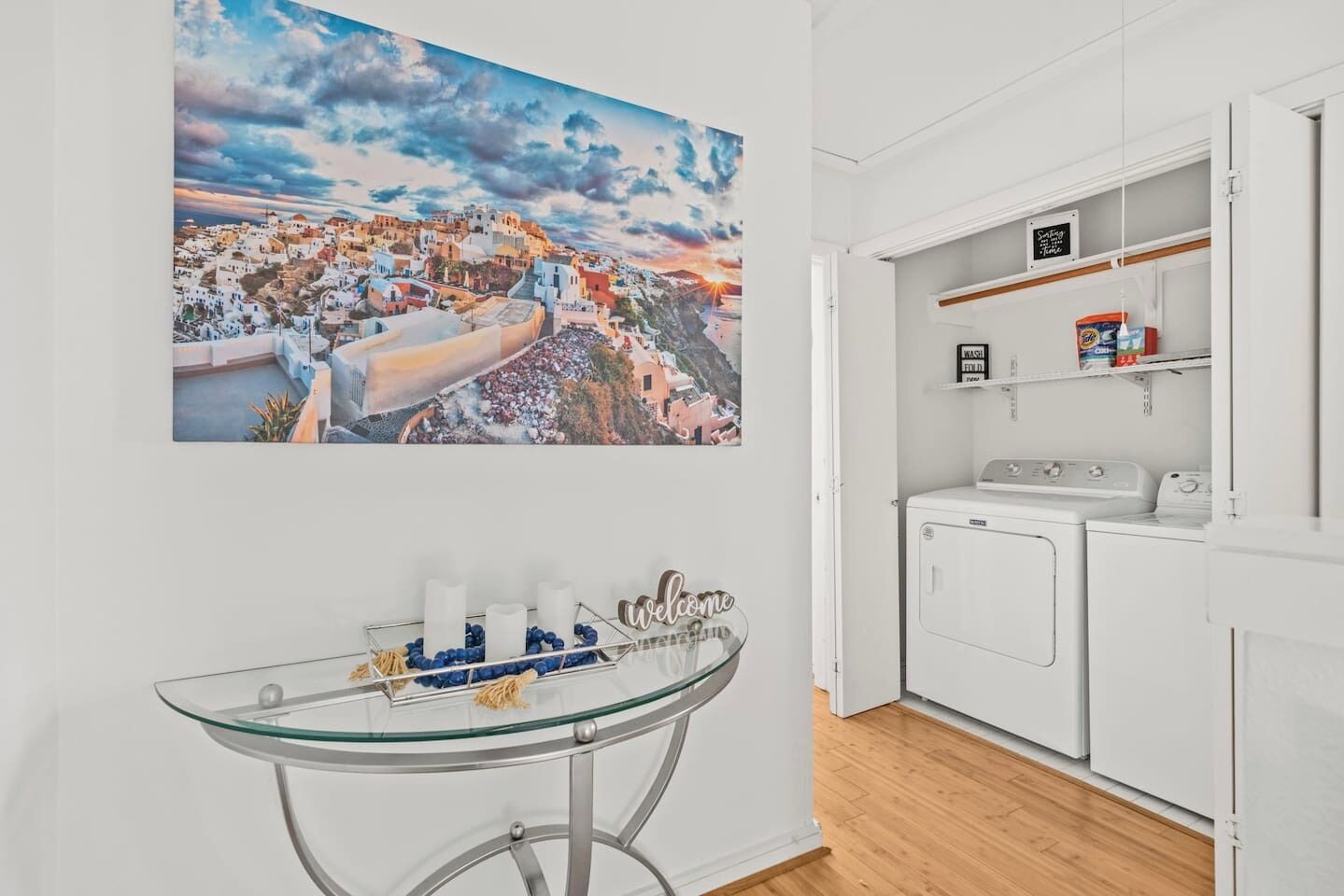 A room with a glass table, a welcome sign, a washing machine, and a large painting of a coastal town.