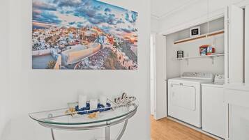 A room with a glass table, a welcome sign, a washing machine, and a large painting of a coastal town.