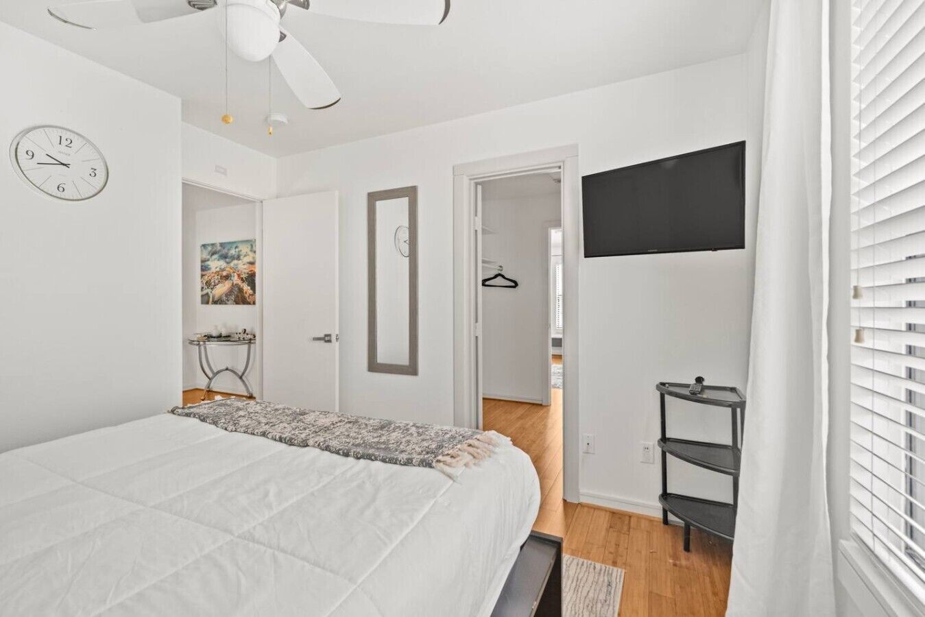 A bedroom with a bed, a ceiling fan, a wall clock, a TV, and a door leading to another room.
