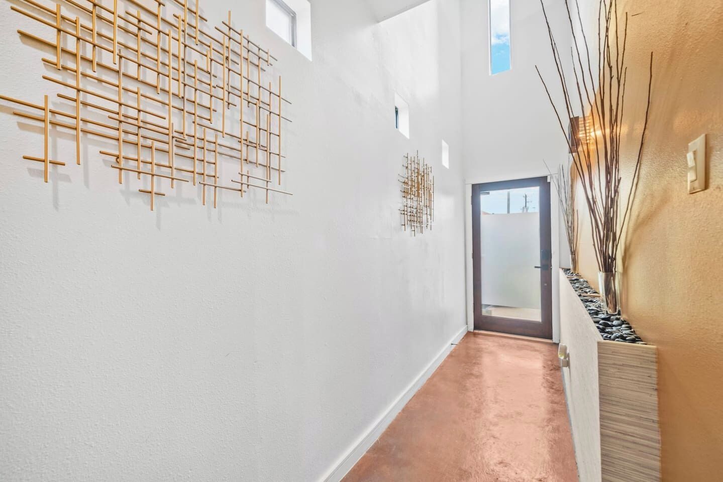 A corridor with a modern design, featuring a wooden art installation on the left wall and a vase with branches on the right.