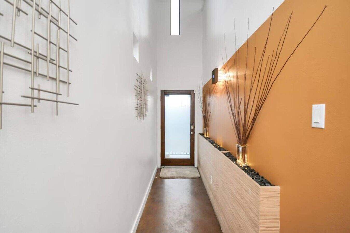 A hallway with a wooden floor, a white wall with a decorative metal rack, and a wooden wall with a lighted plant arrangement.