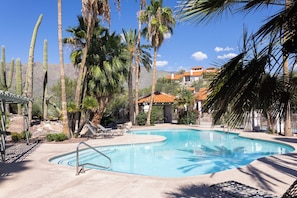 A swimming pool with lounge chairs, palm trees, and a building with a red roof in the background.