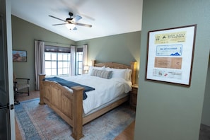 A bedroom with a wooden bed, a ceiling fan, a window with curtains, and a framed event poster on the wall.