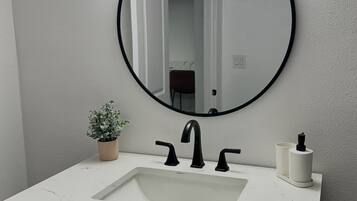 Une salle de bain avec un lavabo en marbre blanc, des robinets noirs, un miroir rond et une petite plante en pot.