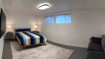 A bedroom with a bed, bedside tables, a flat-screen TV, two windows with blinds, and a gray couch.