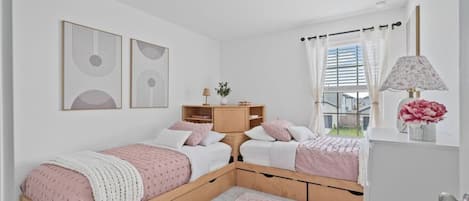 A bedroom with two beds, a bedside table, a nightstand, a window with curtains, and wall art.