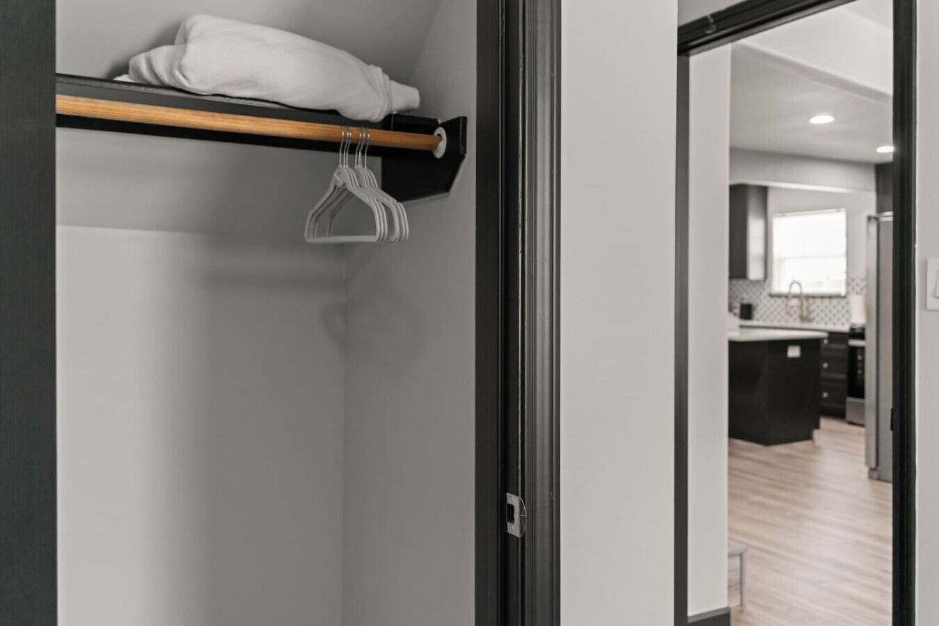 Un interior moderno con un estante de madera que sostiene una toalla blanca y un perchero. Una puerta da acceso a una zona de cocina con suelo de madera.