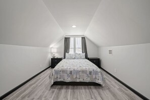 Ein Schlafzimmer mit geneigter Decke, einem Bett mit gemustertem Überwurf, einem Nachttisch mit Lampe und einem Fenster mit Vorhängen.
