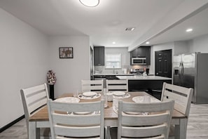 A modern kitchen with dark cabinets, a stainless steel refrigerator, and a dining area with a wooden table and chairs. - Cozy 3-Bedroom Suburban Retreat near Downtown (Houston)