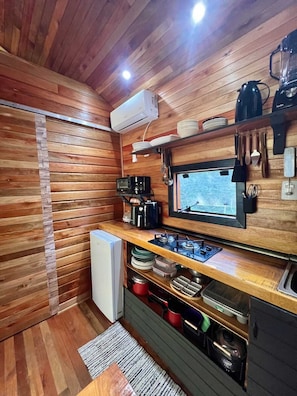 A compact kitchen with wooden walls, a built-in stove, a microwave, and a window with a view. - Frost Cabin • Urubici, SC | Private Stream, your retreat with a hot tub. (Urubici)