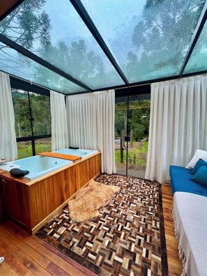 A room with a wooden bathtub, a patterned rug, a blue sofa, and a view of trees through large windows. - Frost Cabin • Urubici, SC | Private Stream, your retreat with a hot tub. (Urubici)