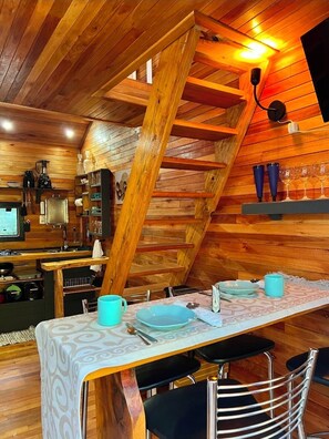 A cozy wooden interior with a dining area featuring a table set for two, a kitchen area with appliances, and a wooden ceiling with exposed beams. - Frost Cabin • Urubici, SC | Private Stream, your retreat with a hot tub. (Urubici)