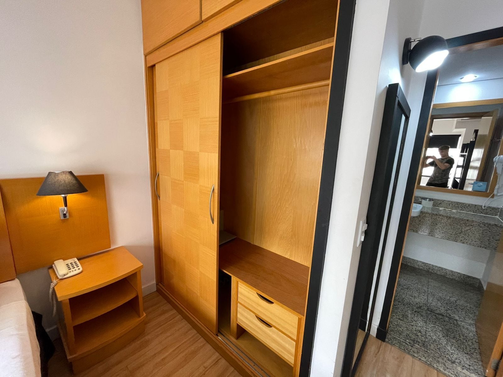 Quarto com guarda-roupa de madeira, mesa de cabeceira com abajur e espelho.